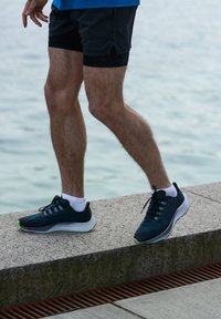 Scarpe da corsa con tomaia in rete traspirante di colore blu navy, dettagli neri, suola verde e intersuola ammortizzata, abbinate a pantaloni corti neri e calze bianche.
