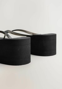 Sorte flade flip-flops med tykke såler og gennemsigtige stropper på en lys baggrund, vist fra bagsiden.