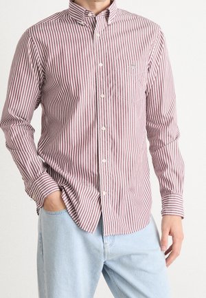 Homme portant une chemise à rayures verticales rouges et blanches à manches longues, boutonnée, avec un jean bleu clair, main dans la poche, debout devant un fond blanc.