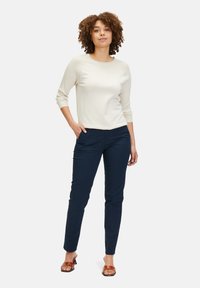 Hellbeige gerippter Pullover mit 3/4-Ärmeln, kombiniert mit taillierten, navyblauen Hosen. Das Model steht mit den Händen in den Taschen. Braune Sandalen mit Riemen.