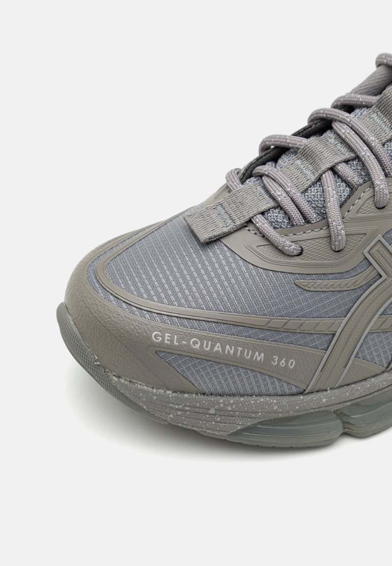 Grauer Sportschuh mit einer strukturierten Oberseite, die ein Mesh-Design aufweist, Schnürsenkel und eine Gel-Dämpfungsmittelsohle. Auffällige Markierung an der Seite.