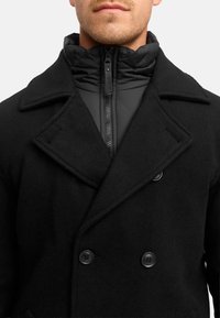 Schwarzer doppelt knöpfbarer Mantel aus Wolle, mit Schalkragen, großen Knöpfen und einer gesteppte Innjacke mit hohem Kragen.