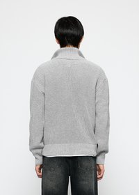 Person med kort svart hår iført en lys grå strikkegenser og falmede svarte jeans, sett bakfra mot en nøytral bakgrunn.