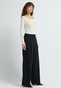 Crème lange mouwen top met een off-shoulder halslijn, gecombineerd met zwarte wijde broek. De broek heeft een gladde textuur met voorplooien.