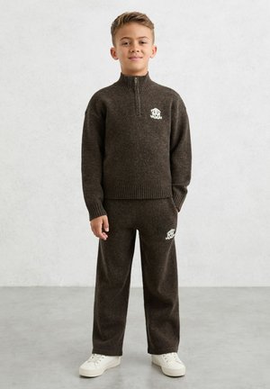 Brun strikket sweater med høj krave og hvidt logo; matchende brune bukser; dreng iført hvide sneakers; blød tekstur.