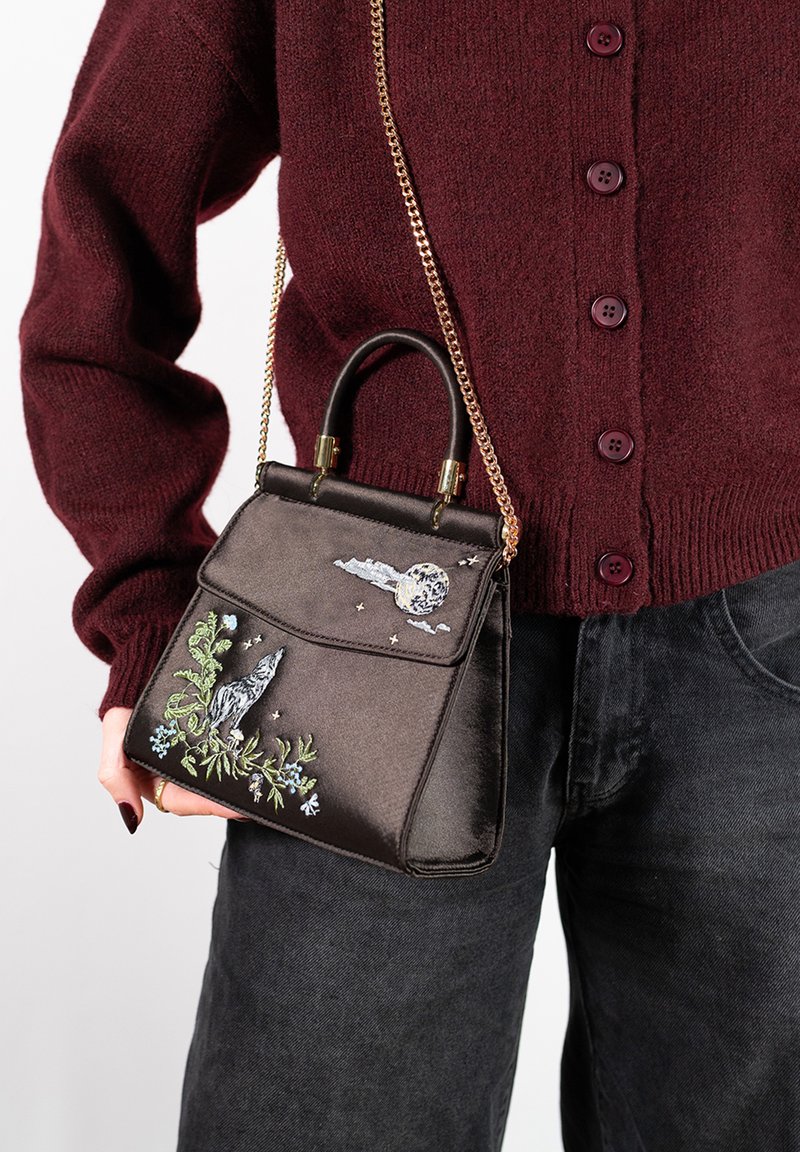 Sac à main en satin marron avec broderie d'un loup et d'éléments célestes. Il présente une forme structurée, une poignée supérieure et une longue bandoulière en chaîne dorée.