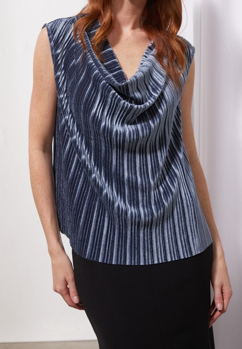 Haut sans manches en velours bleu avec des plis verticaux et un col bénitier drapé porté avec une jupe noire.