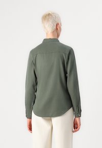 Personne aux cheveux blonds courts vêtue d'une chemise à manches longues vert olive et d'un pantalon écru, vue de dos sur un fond uni.