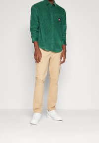 Zelená manšestrová košile na knoflíky s náprsní kapsou, kombinovaná s béžovými cargo kalhotami a bílými teniskami.