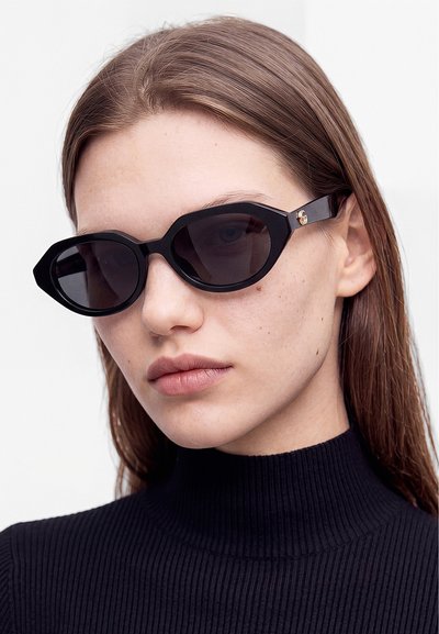 Femme aux cheveux bruns et raides, portant des lunettes de soleil noires géométriques et un pull à col roulé noir côtelé, sur un fond uni.