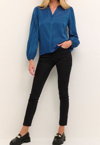 Blå satinbluse med V-udskæring, lange ballonærmer og en afslappet pasform, parret med sorte skinny jeans og sorte loafers.