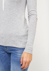 Primo piano di una persona che indossa una camicia a maniche lunghe grigia chiaro e jeans blu, mostrando il braccio destro e la mano rilassati lungo il fianco.
