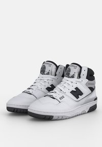 Białe sneakersy za kostkę z czarnymi akcentami, cholewka ze skóry i materiałów syntetycznych, wyściełany kołnierz, teksturowana podeszwa oraz wyraźne logo "N" po bokach.