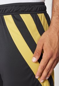 Schwarze Sportshorts mit gelben diagonalen Streifen; elastischer Bund und glattes synthetisches Material; Hand, die an der Seite aufliegt und die Textur zeigt.
