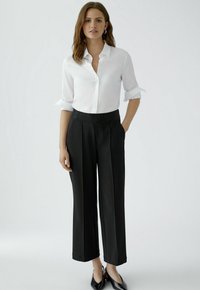 Weißes Hemd mit aufgekrempelten Ärmeln, kombiniert mit schwarzen, weit geschnittenen Hosen mit strukturiertem Taillenbund und dezenten Falten. Schwarze spitz zulaufende Schuhe.