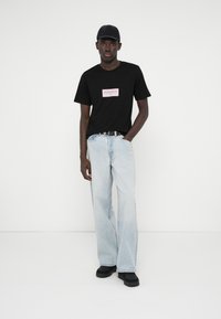 Μαύρη μπλούζα με λογότυπο σε άσπρο και κόκκινο, ανοιχτό μπλε φαρδύ τζιν παντελόνι, μαύρη ζώνη και μαύρα αθλητικά παπούτσια; το μοντέλο φοράει μαύρο καπέλο.