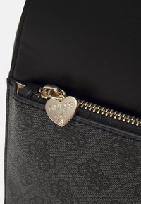 Sac en cuir noir texturé avec quincaillerie dorée, doté d'un tirette de fermeture éclair en forme de cœur gravée "GUESS girl" et d'un accent en pierre.