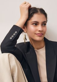 Svart kavaj med knappade ärmslutar, beige ribbad topp och guldfärgade hängande örhängen med metallremsor i olika längder. Slät hud och ljust hår.