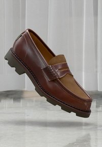 Brown leather loafer with suede panel and chunky rubber sole, floating above a marble floor against a white vertical panel background.
