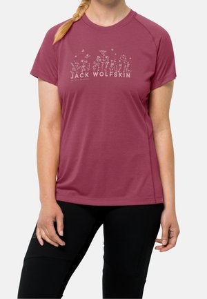 Kvinde iført en kortærmet vinrød skjorte med vilde blomster-illustrationer og "Jack Wolfskin"-logo, kombineret med sorte bukser.