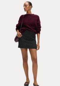 Burgunderfarbener Strickpullover mit lockerer Passform. Schwarzer Minirock mit einem weißen Kreuzmuster. Das Modell hat kleine Ohrringe und eine Tasche als Accessoires.
