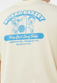 Twee cartoonfiguren houden een surfboard vast met de tekst "Rip Curl Surf Shop" en contactgegevens op de achterkant van een crèmekleurig shirt.