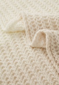 Textile tricoté de couleur crème avec un tissage lâche texturé et une finition douce et duveteuse. Les bords présentent une section repliée pour le détail.