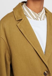 Blazer de pana color caqui con solapas de muesca y cierre de un solo botón, superpuesto a una camiseta blanca de cuello redondo y una camisa de rayas con cuello.