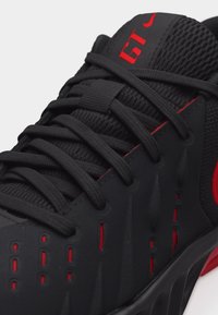 Zbliżenie na czarny i czerwony sportowy sneaker z detalicznym sznurowaniem, oddychającą siateczką oraz logo "GT" na języku.