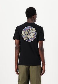 Tricou negru cu mânecă scurtă, având un grafic circular pe spate cu "SANTA CRUZ" scris într-un font stilizat și cu modele geometrice colorate.