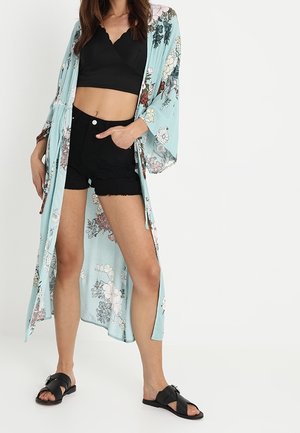Vrouw draagt een zwart cropped topje, zwarte shorts met hoge taille, lichtblauw bloemenkimono en zwarte slip-ons, staand met hand in zak.