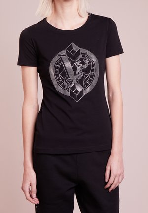 Femme portant un t-shirt noir ajusté avec un motif géométrique et animalier blanc sur le devant, associé à un pantalon noir, debout devant un fond neutre.