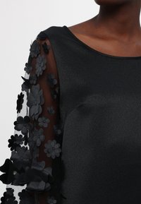 Robe noire avec manches longues transparentes ornées d'applications florales noires en 3D sur une personne à la peau foncée.