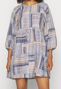 Leichtes, gemustertes Kleid aus einer Mischung von blauen und beigen Stoffen, mit rundem Ausschnitt und 3/4-langen Puffärmeln. Mit Taschen.