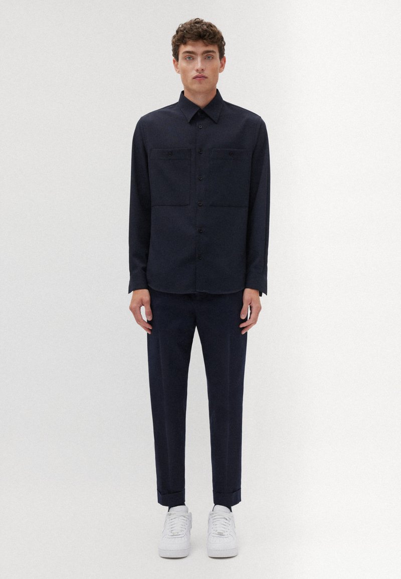 Mann, der vor einem weißen Hintergrund steht, trägt ein dunkelblaues Hemd mit Knöpfen und Taschen, passende Hosen, die an den Knöcheln umgeschlagen sind, und weiße Sneaker.