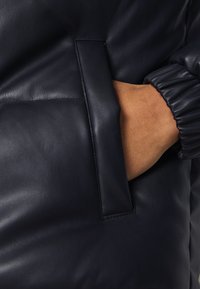 Main dans la poche d'une veste en cuir noire avec un poignet élastique et une ouverture de poche cousue sur la veste.