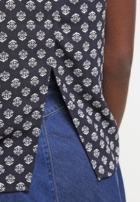 Top de tela azul oscuro con patrones geométricos blancos, que presenta una abertura lateral y una textura suave, combinado con material de mezclilla.