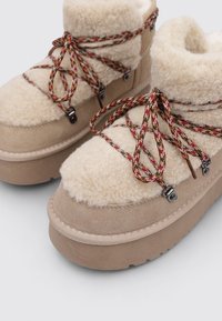 Bottes en daim beige avec des dessus en fourrure crème, lacets à motifs multicolores et ferrures métalliques robustes, semelles compensées texturées.