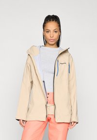 Μπεζ αδιάβροχο μπουφάν με ανοιχτό μπλε εσωτερικό, ρυθμιζόμενη κουκούλα και τσέπες με φερμουάρ. Φορεμένο πάνω από γκρι μπλούζα και πορτοκαλί παντελόνι.