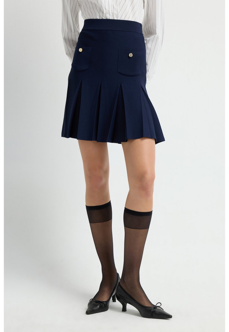 Falda plisada azul marino con dos bolsillos frontales y acentos de botones dorados, combinada con medias negras transparentes hasta la rodilla y tacones negros de punta. 