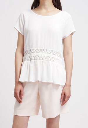 Femme portant un chemisier blanc à manches courtes avec des bandes en dentelle à la taille et un short rose clair arrivant aux genoux.