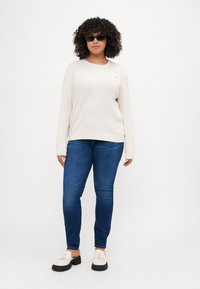 Kremfarget strikket genser med teksturert mønster, blå tettsittende jeans og hvite sko med frynse-detaljer. Modell med svarte solbriller.
