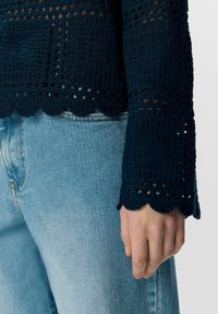 Pull en crochet bleu marine à manches longues, avec des poignets en dentelle et un design en tricot ouvert. Associé à un jean en denim bleu clair.