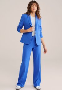 Svetlomodrý oblek s bundou a nohavicami z hladkého materiálu, v kombinácii so sivou košeľou. Bunda má jeden gombík a úzky strih. Mäkká textúra.