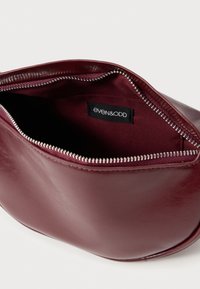 Πορτοκαλί τσάντα από συνθετικό δέρμα μπορντό με πάνω φερμουάρ, εσωτερική φόδρα και εμφανές λογότυπο της μάρκας. Λείο υφή και στρογγυλεμένο σχήμα.