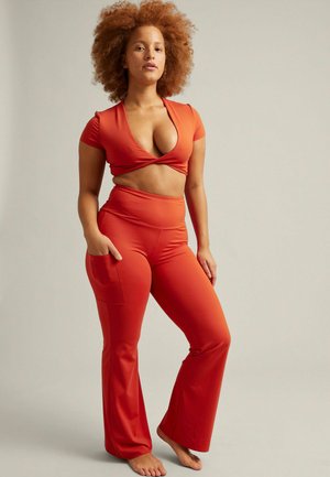 Vrouw met krullend haar staat blootsvoets, draagt een rood crop top en hoge wijde broek met één hand in de zak, tegen een effen achtergrond.