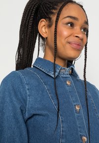 Camisa de ganga azul com frente com botão, colarinho clássico e costuras distintas. O tecido tem uma aparência suave e texturada.