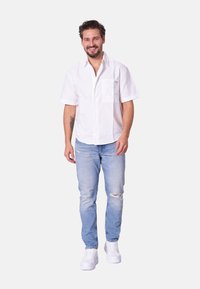 Camisa blanca de manga corta con botones y un bolsillo, combinada con jeans claros rasgados y zapatillas blancas. Textura de tela suave.