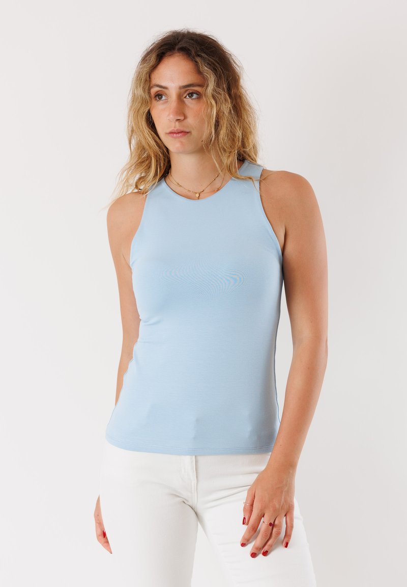 Top senza maniche blu chiaro realizzato in tessuto morbido, con un design aderente e un collo alto. Indossato con pantaloni bianchi.