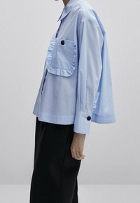 Chemise bleu clair à boutons avec une poche à volants, des boutons noirs en accent, une coupe oversize et des manches retroussées. Portée avec un pantalon foncé.